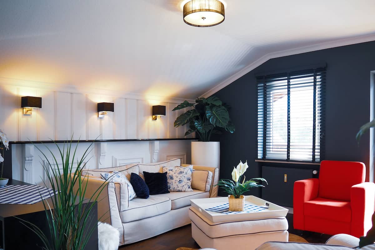 Una sala de estar moderna con un sofá blanco, un sillón rojo y una mesita de centro. Hay luces fijadas en la pared y una planta grande.