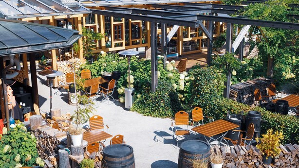 Un área de estar al aire libre con mesas y sillas de madera, rodeada de vegetación y una estructura de pérgola.