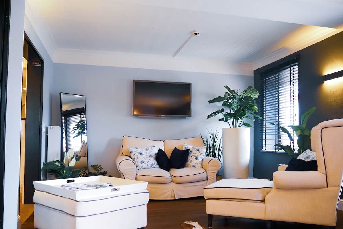 Una sala de estar moderna con sofá, sillón y reposapiés, televisor de pantalla plana y ventanas amplias con persianas.