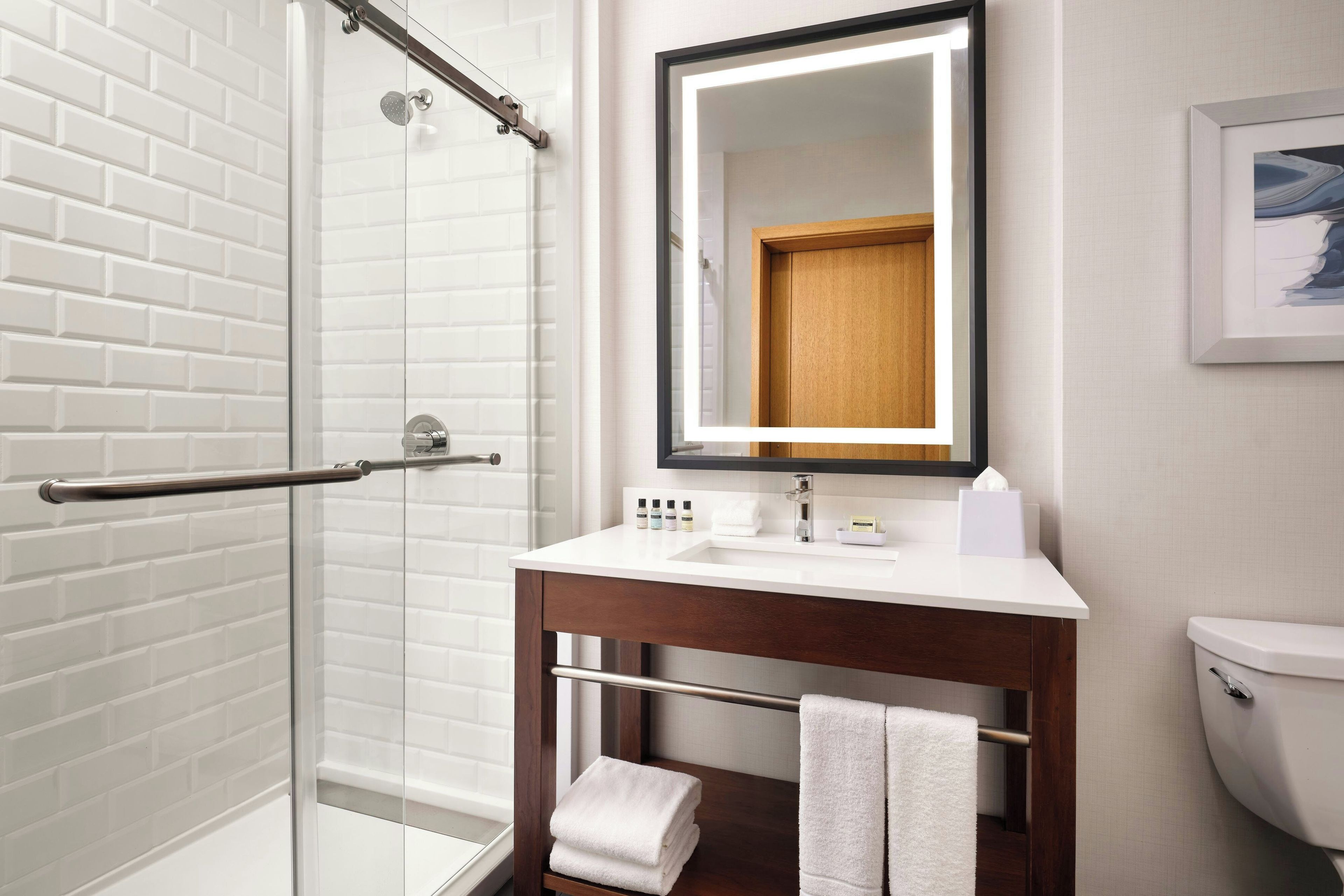 A bathroom with a glass shower enclosure, a white sink, a wooden vanity, and a framed picture on the wall.
