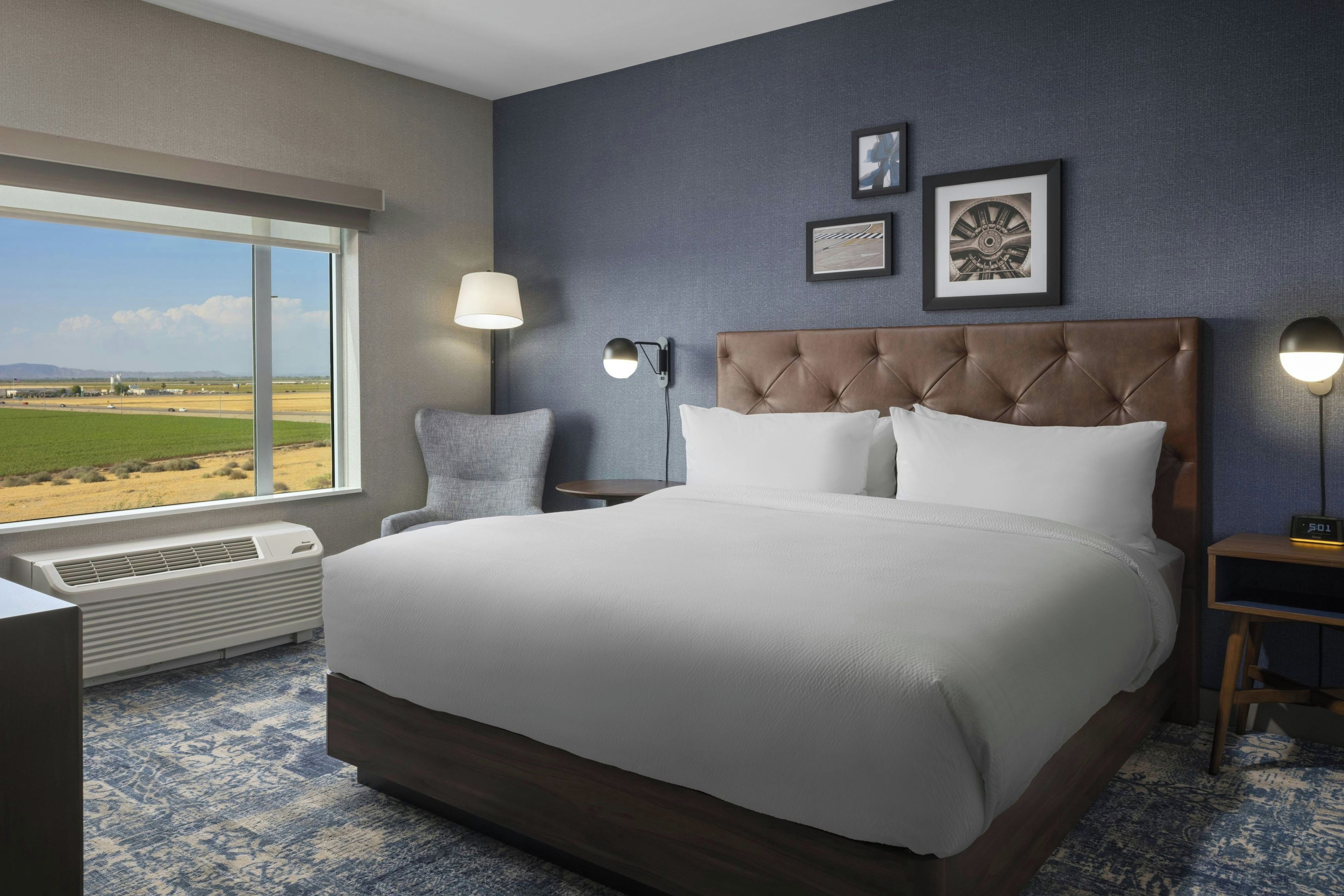 A hotel room with a large bed, a chair, a desk, a window with a view of the countryside, and two wall-mounted lamps.