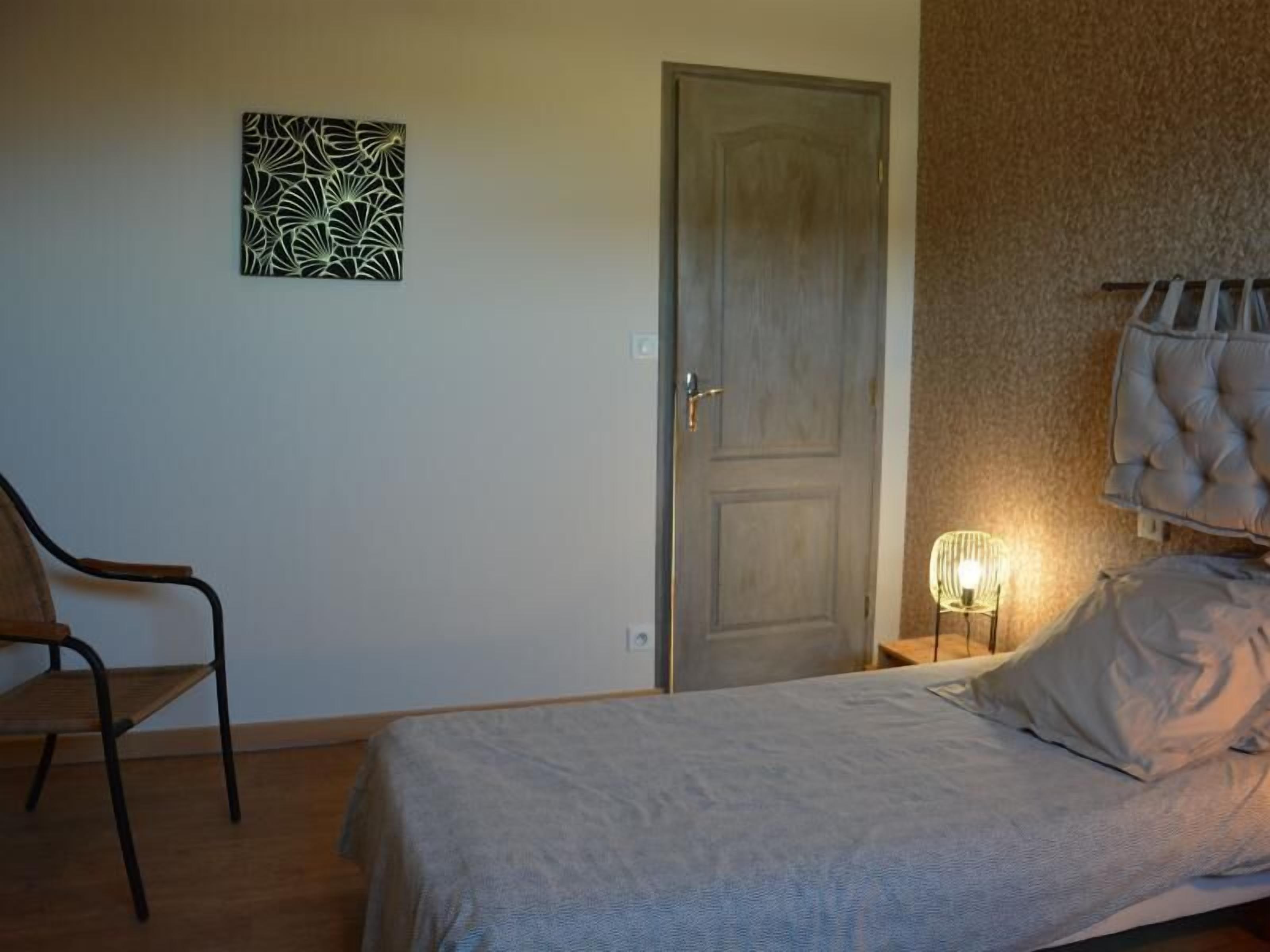 A bedroom with a bed, a chair, a door, and a wall-mounted light fixture.