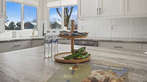 A kitchen with white cabinets, a marble countertop, a water dispenser, a tray with snacks, and a brochure on the counter. - Modern Farmhouse Retreat | 5BR | With Pool Views (Meridian)