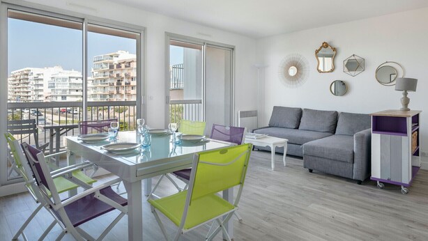 Een modern appartement met een balkon, een eethoek met een glazen tafel, een grijze bank en een kleine witte tafel.