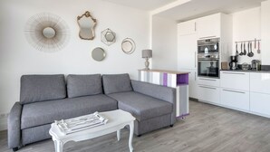 Een moderne woonkamer met een grijze bank, een witte salontafel en een keuken met witte kasten en apparatuur.