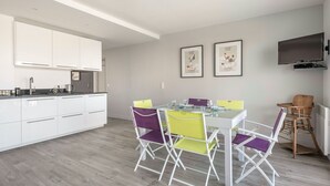 Een moderne keuken met witte kasten en een eethoek met kleurrijke stoelen. Twee ingelijste foto’s hangen aan de muur en er is een tv boven de eettafel bevestigd.