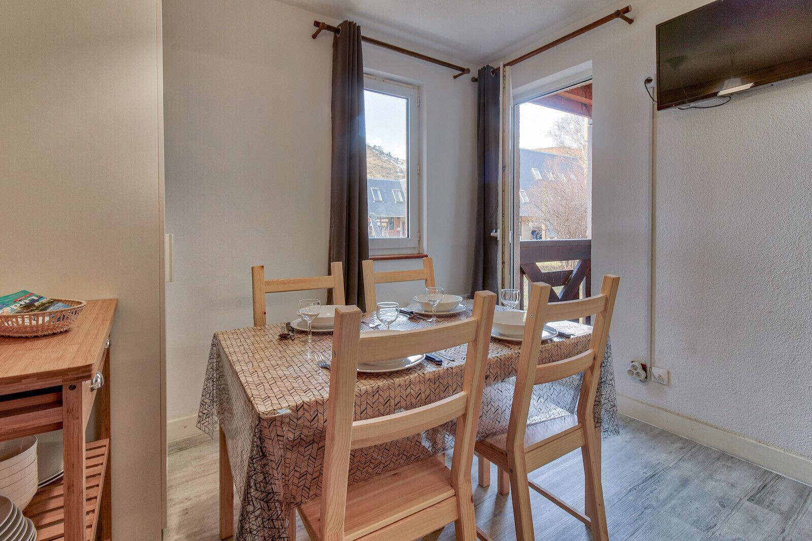 A dining area with a table set for four, wooden chairs, a basket on a side table, and a view through a window.