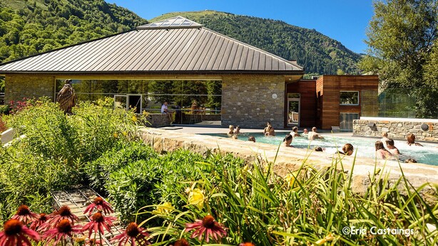 A pool area with people swimming, surrounded by lush greenery and a stone building with a sloped roof.