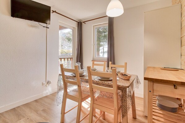 A dining area with a wooden table and chairs, a television mounted on the wall, and a window with curtains. - Direct access to the slopes - flat for 4 (Genos)