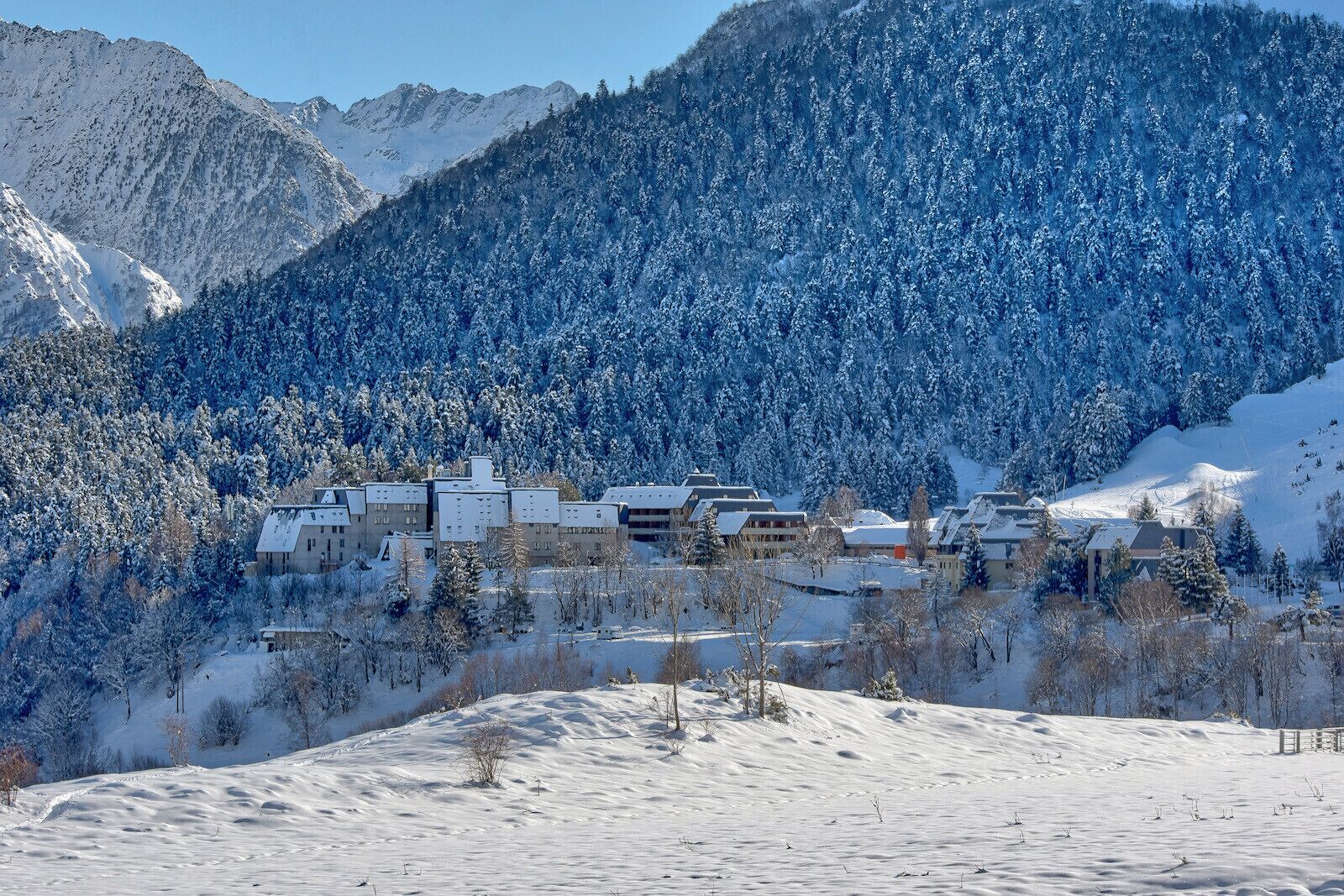 Un village de montagne enneigé, avec des bâtiments regroupés, entouré de forêts denses et de pentes recouvertes de neige.