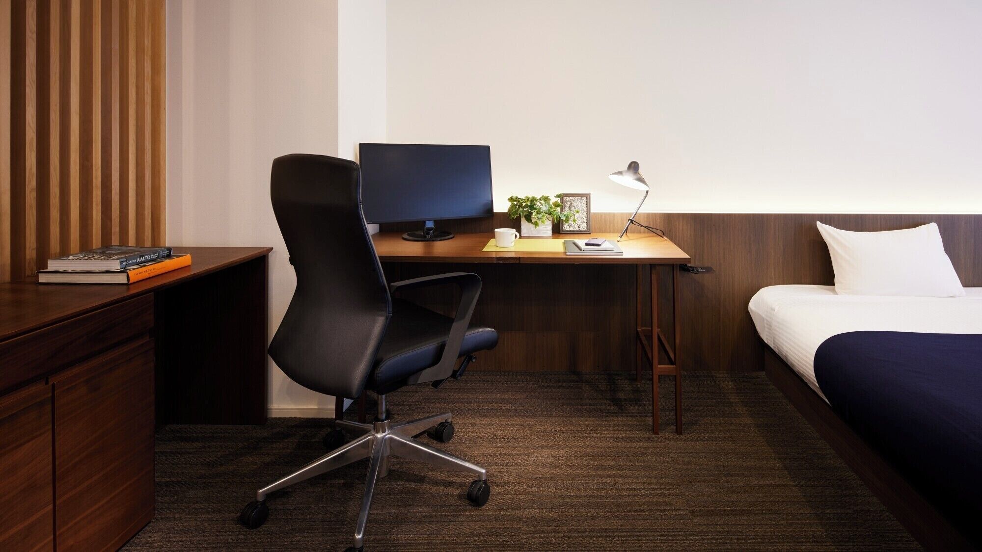 A hotel room with a wooden desk, a black office chair, a bed with white bedding, and a small plant on the desk.