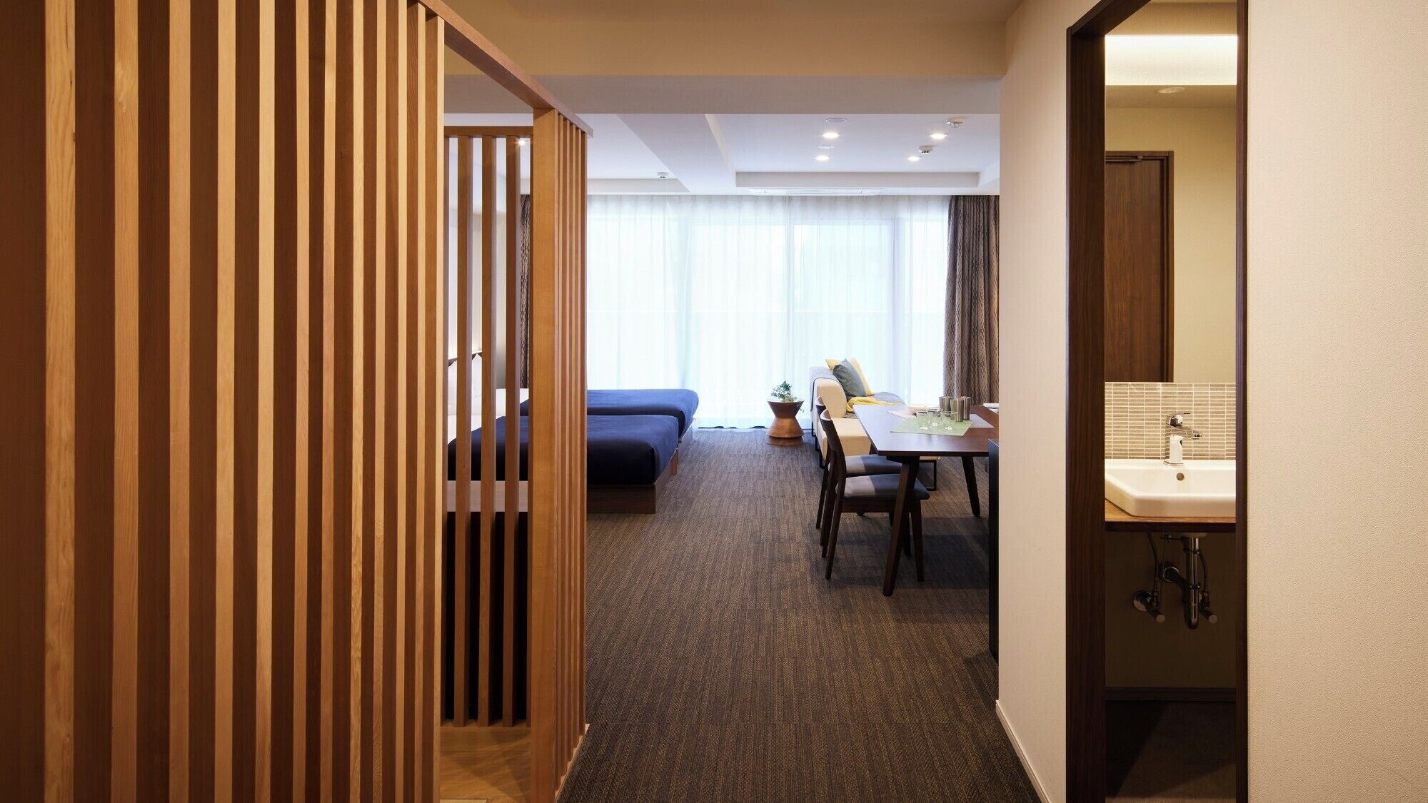 A hotel room with a bed, a sofa, a dining table with chairs, and a bathroom with a sink and mirror.
