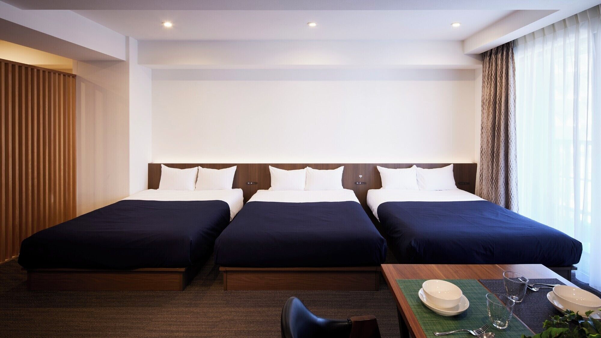 Een hotelkamer met drie bedden, een houten hoofdbord, een eettafel met een hoed en een bril, en een raam met gordijnen.