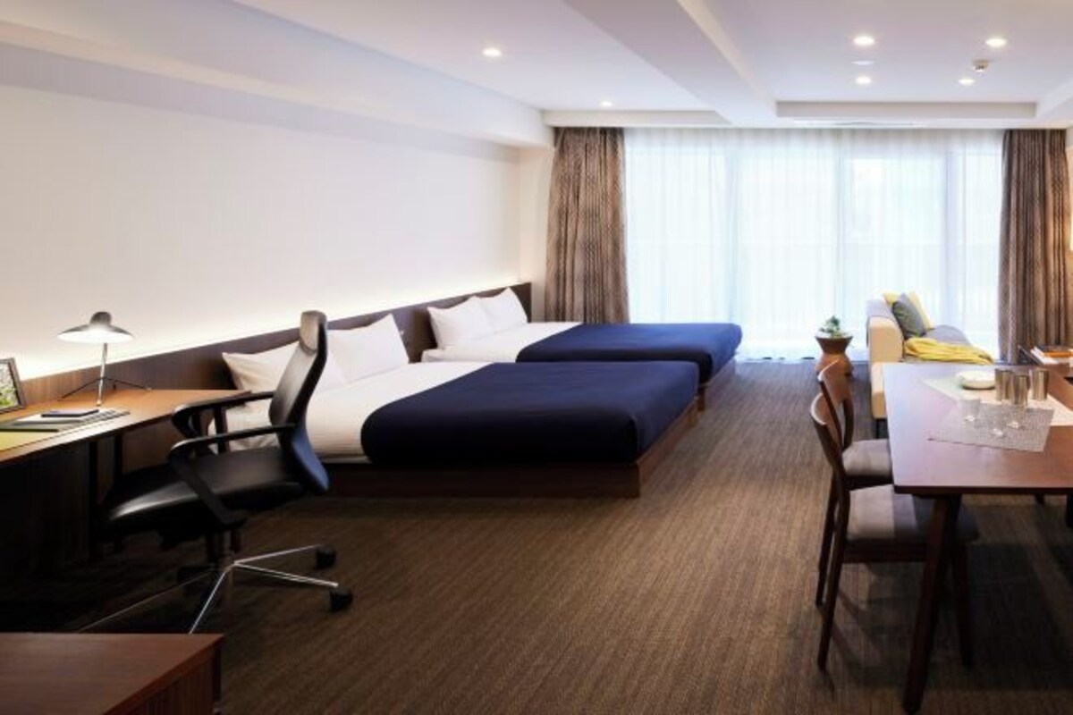 A hotel room with two beds, a desk, a chair, a dining table with chairs, and a window with curtains.