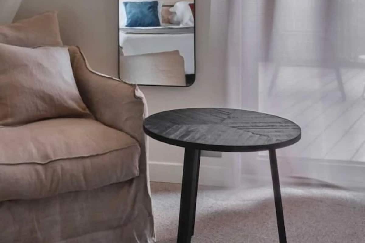 A modern room with a beige sofa, a round black table, and a large mirror reflecting a bed with pillows.