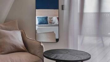 A modern room with a beige sofa, a round black table, and a large mirror reflecting a bed with pillows.