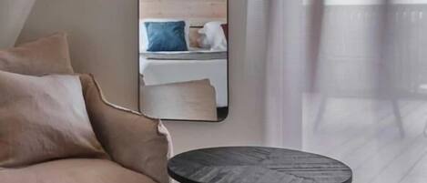 A modern room with a beige sofa, a round black table, and a large mirror reflecting a bed with pillows.