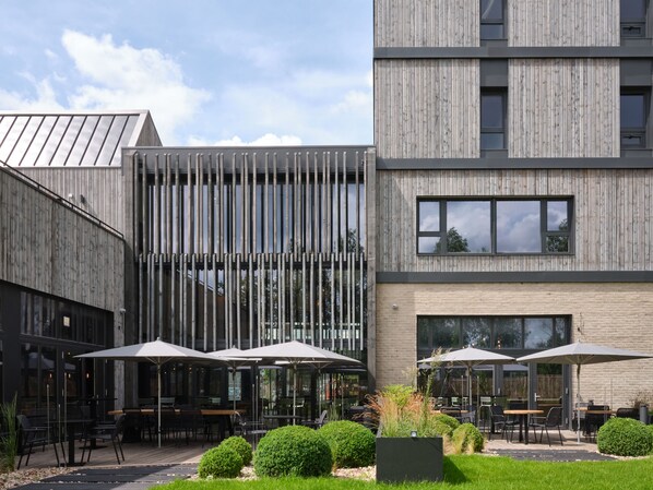 A modern building with a wooden facade, outdoor seating area with umbrellas, and a well-maintained garden. - Bercail Hotel (Wambrechies)