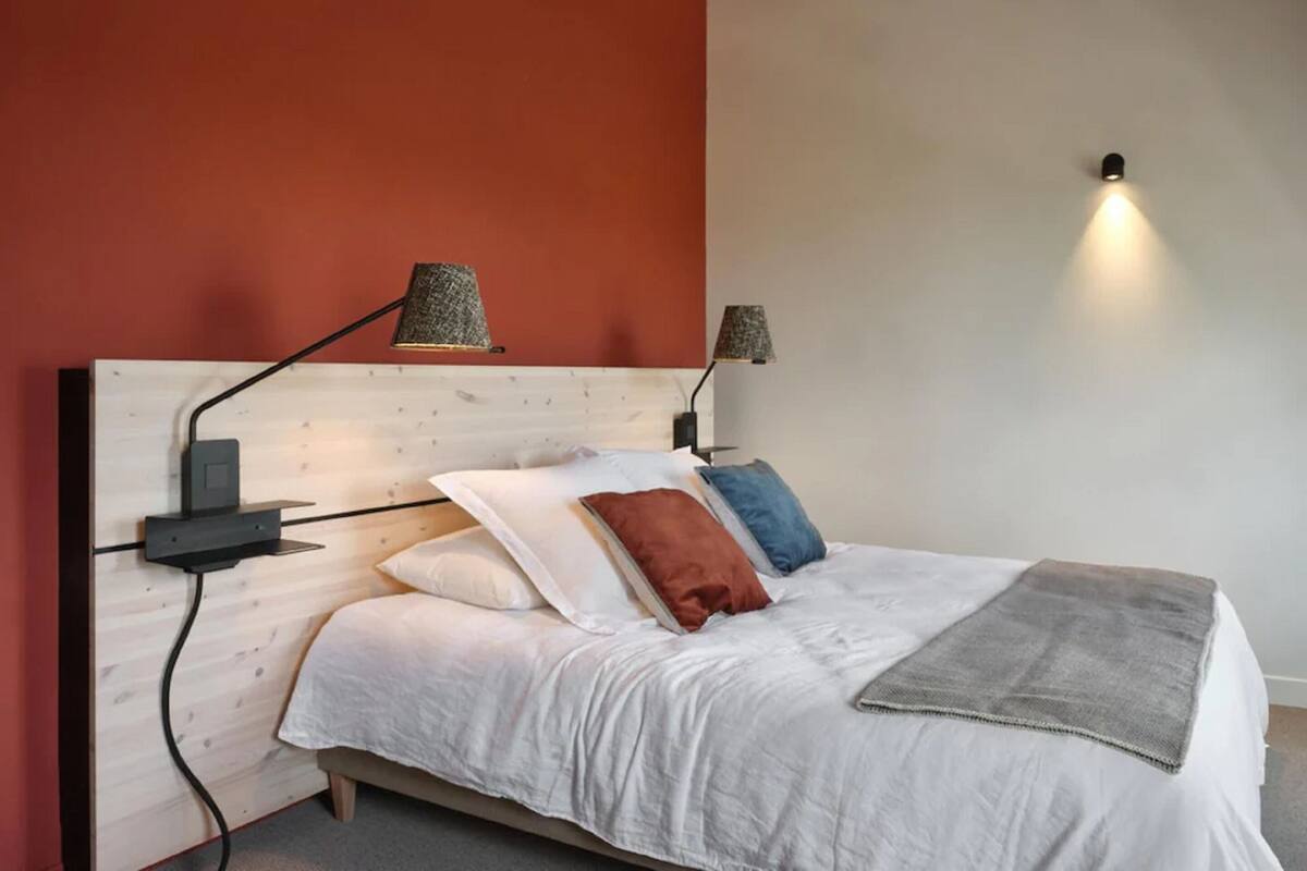 A bedroom with a wooden headboard, a white bedspread, and multiple pillows. There are two wall-mounted lamps and a bedside table with a lamp.