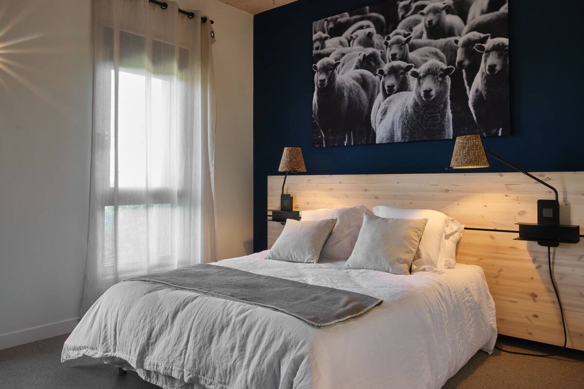 A bedroom with a bed, white bedding, a headboard with a wooden panel, two wall-mounted lamps, and a large black-and-white photograph of sheep on the wall.