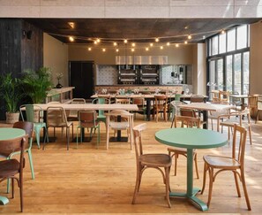A spacious dining area with multiple tables and chairs, a kitchen in the background, and large windows allowing natural light. - Bercail Hotel (Wambrechies)