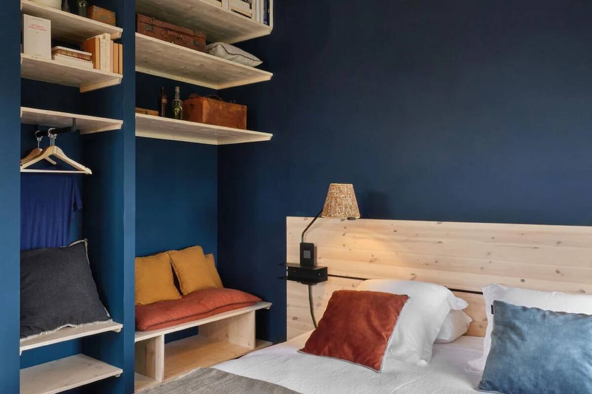 A bedroom with a bed, a wooden headboard, a bedside table with a lamp, and built-in shelves with books and a hanging garment.