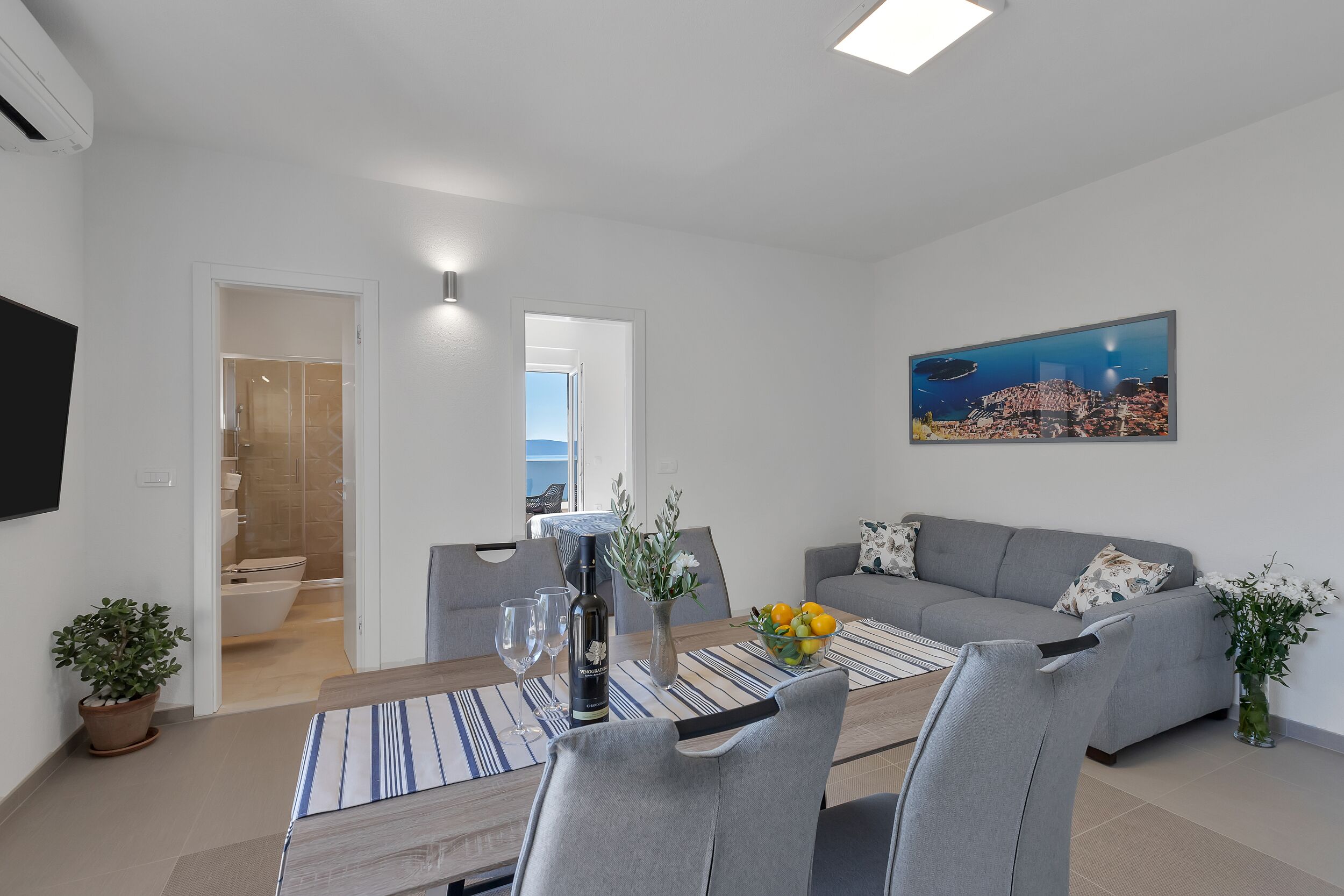 A modern living room with a dining area, grey sofas, a flat-screen TV, and a painting of a cityscape.