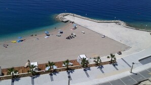 An aerial view of a beach with clear blue water, a sandy area with umbrellas and lounge chairs, and a wooden deck with palm trees. - Luxury Apartment Podgora-Zora (Podgora)