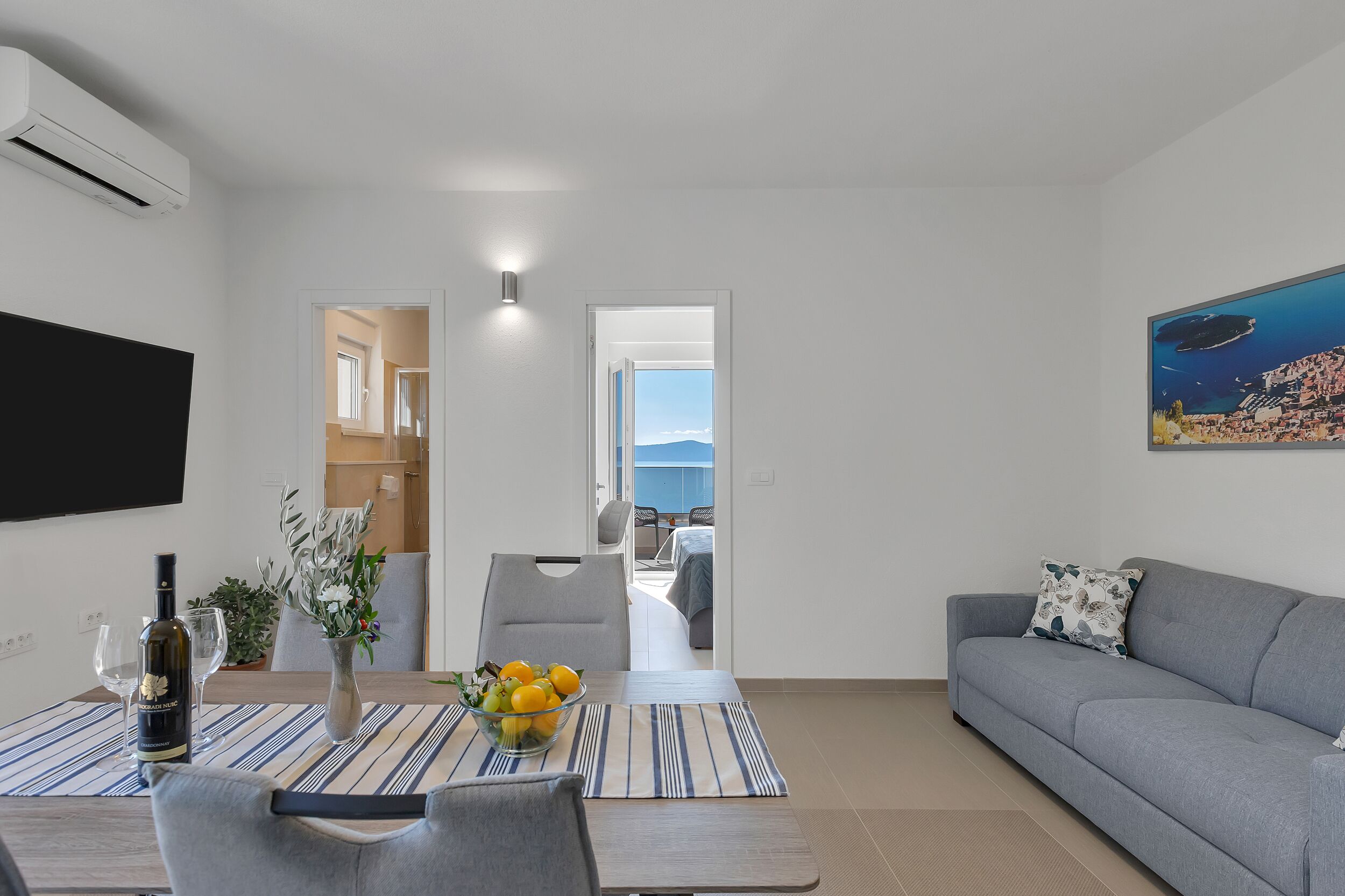 A modern living room with a sofa, a dining table with a vase of flowers, and a TV mounted on the wall.