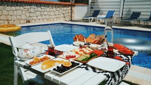 Mesa junto a la piscina con diversos alimentos y bebidas, rodeada de sillones de descanso y una pared de piedra.