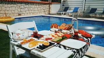 Mesa junto a la piscina con diversos alimentos y bebidas, rodeada de sillones de descanso y una pared de piedra.