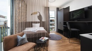 Habitació d'hotel moderna amb un llit, un sofà, una taula petita amb una revista, un escriptori amb una cadira i una televisió penjada a la paret.