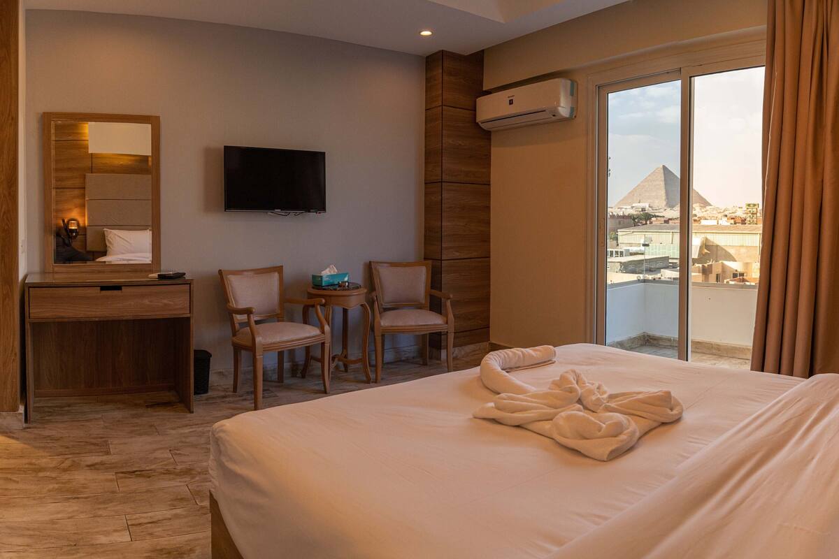 Habitación de hotel con una cama grande, un escritorio, dos sillas, un televisor y vista a una pirámide a través de un ventanal amplio.