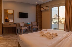 A hotel room with a large bed, a desk, two chairs, a television, and a view of a pyramid through a large window. - Nine Pyramids View Hotel (Giza)