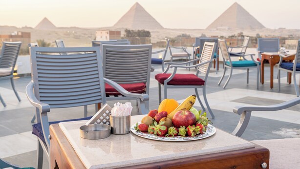 A table with fruits and a plate of food, surrounded by lounge chairs with cushions, overlooking pyramids. - Nine Pyramids View Hotel (Giza)