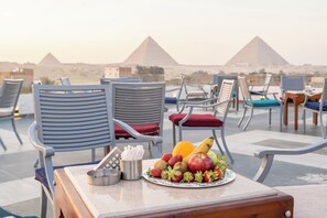 A table with fruits and a plate of food, surrounded by lounge chairs with cushions, overlooking pyramids. - Nine Pyramids View Hotel (Giza)
