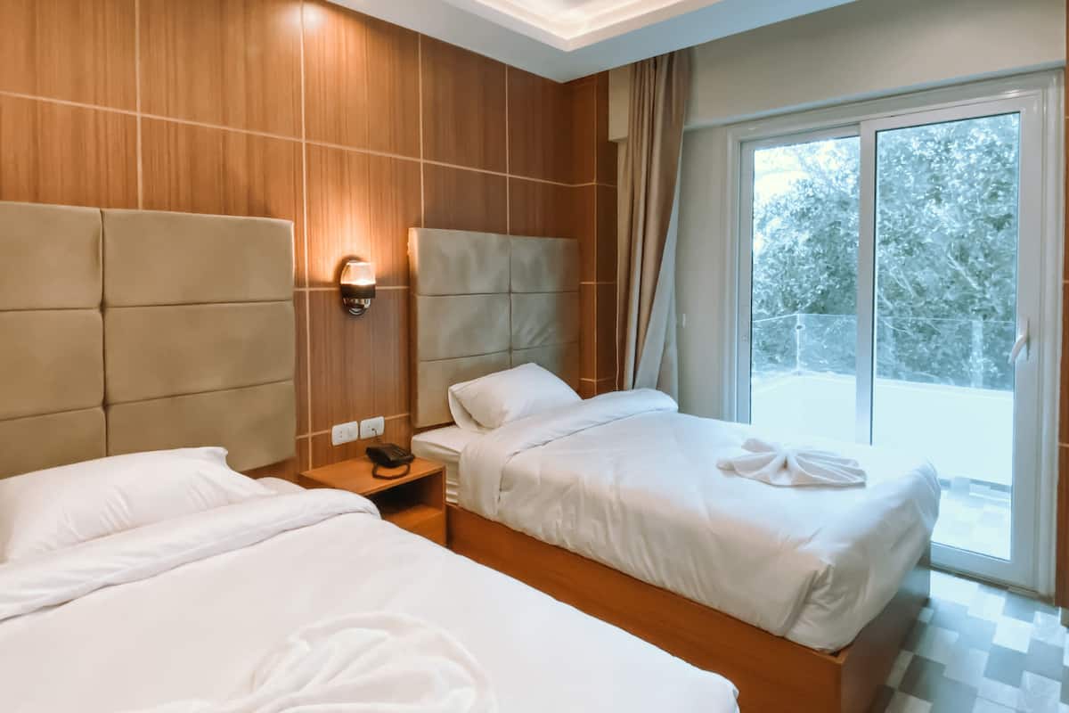 Habitación de hotel con dos camas, cabecera de madera, mesita de noche con teléfono y un ventanal con vistas a árboles.