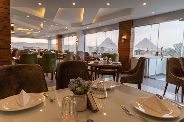 A restaurant with tables set for dining, large windows offering a view of a pyramid, and a modern interior design. - Nine Pyramids View Hotel (Giza)