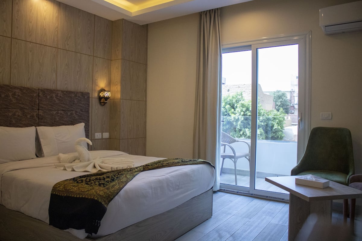Habitación de hotel con una cama grande, un sillón verde, una mesita y un balcón con puerta de vidrio corredera.