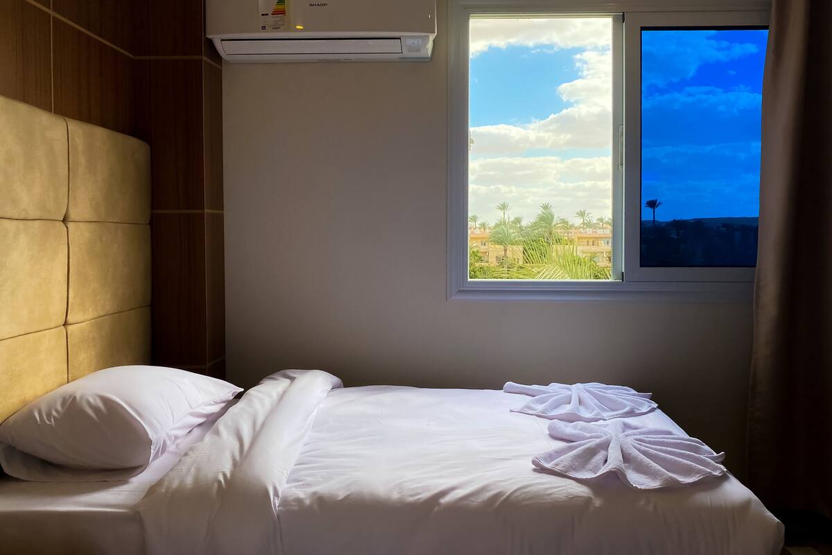 Una cama bien hecha con sábanas blancas y dos toallas, un cabecero de madera, una ventana con vistas a palmeras y edificios, y un equipo de aire acondicionado sobre la cama.