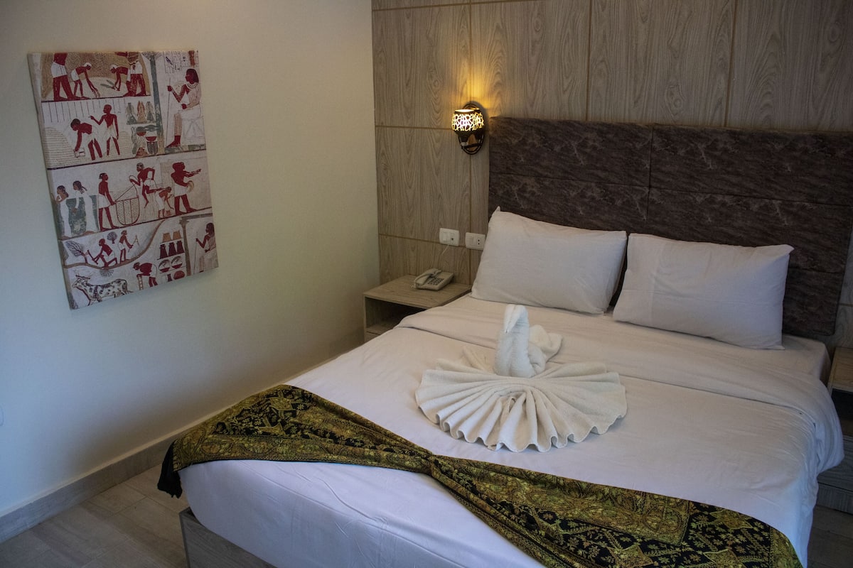 Una cama bien hecha con una escultura de toalla, una lámpara de pared y una mesita de noche pequeña con un teléfono.