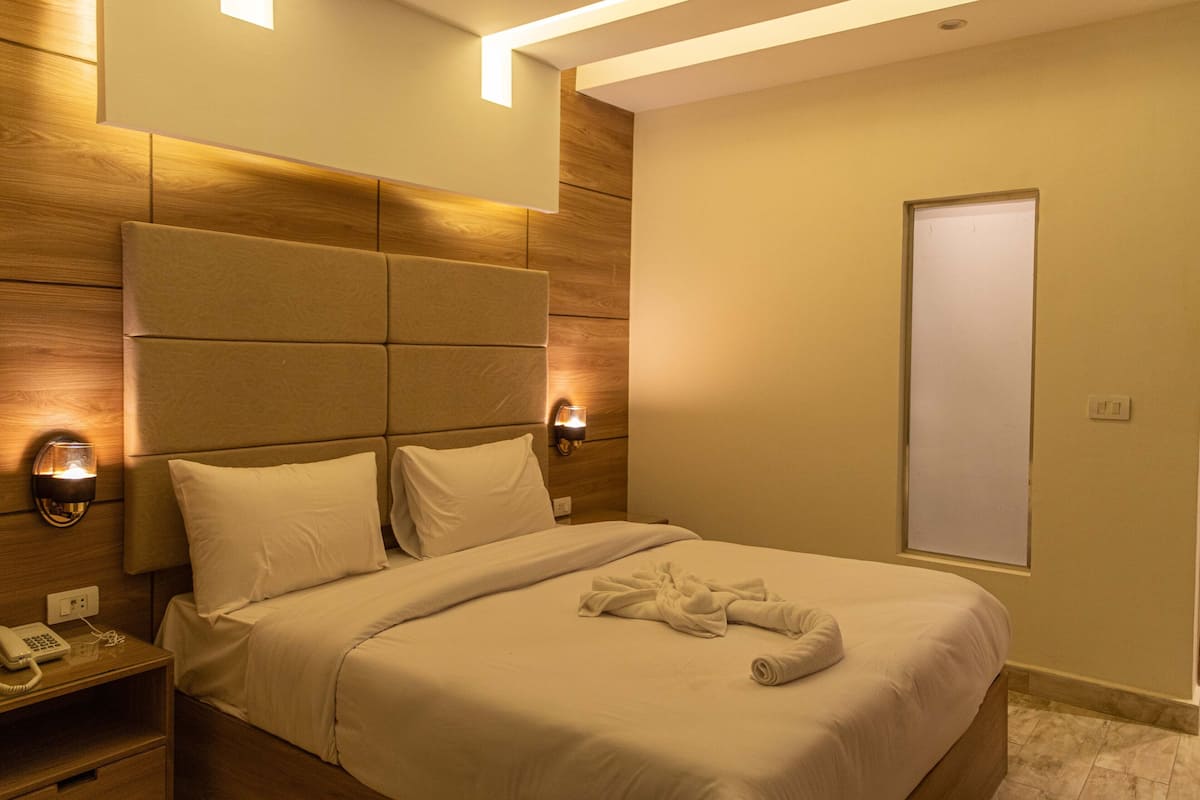 Una habitación de hotel con una cama grande, dos lámparas de noche, un cabecero de madera, una mesita de noche con teléfono y una ventana con persianas.