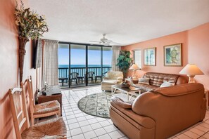 A living room with a sofa, chairs, a TV, and a balcony with a view of the ocean.