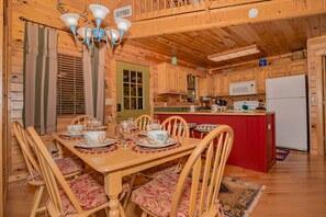A wooden cabin with a dining area featuring a table set for four, wooden chairs, a kitchen with a microwave and refrigerator, and a red island counter.