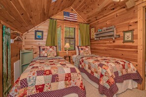 A cabin bedroom with two beds, a window with green curtains, a wooden ceiling, and a wall-mounted shelf with decorations.