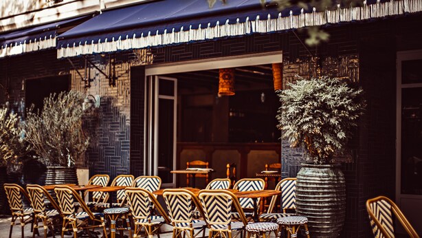 An outdoor cafe with wicker chairs and tables, a large potted plant, and a blue awning. - Babel Belleville (Paris)