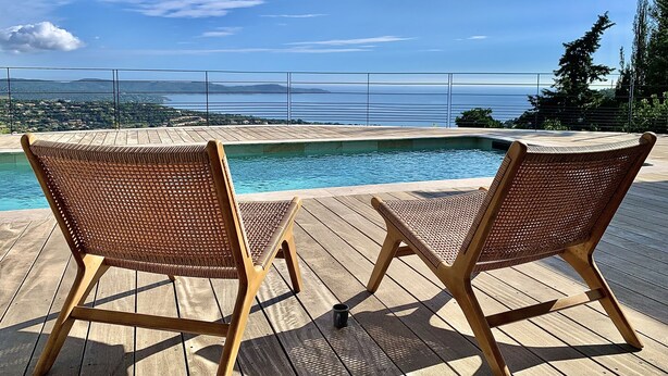 Eine Holzterrasse mit zwei Korbstühlen, die einen Pool und einen weiten Blick auf das Meer und das Umland bieten.