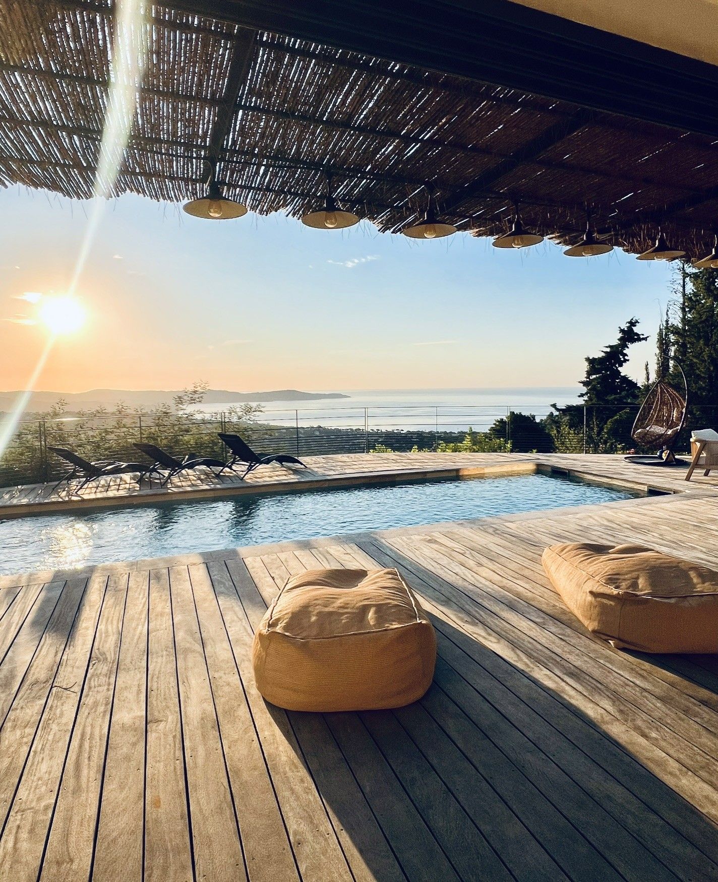 Een houten terras met een zwembad, twee hangzakken en uitzicht op zee bij zonsondergang.