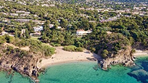Vista aérea de una zona costera con playa, aguas turquesas cristalinas y una zona residencial densamente poblada con casas y vegetación.