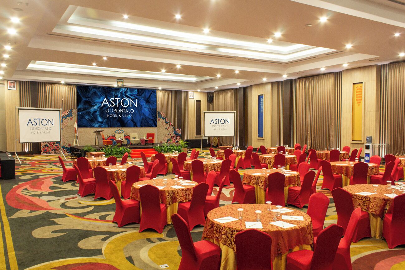 Une salle de conférence avec des tables rondes et des chaises rouges, un grand écran affichant le texte « ASTON GORONTALO HOTEL & VILLAS », et une scène avec un pupitre.